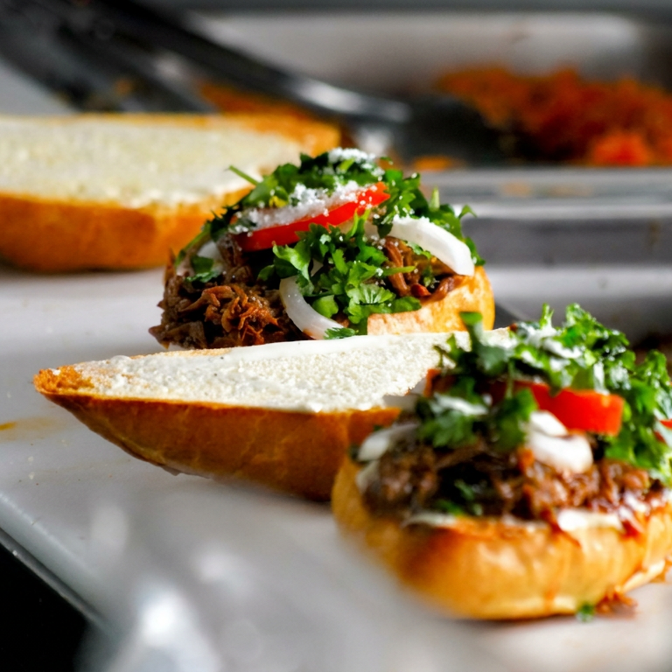 Open sandwiches filled with shredded meat, fresh herbs, tomatoes, onions, and sauce on a restaurant prep counter.